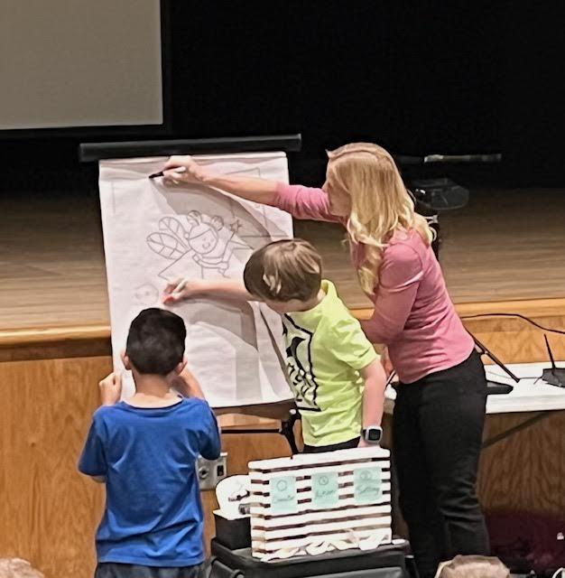 Eli Juarez (grade 3) and Tyler Frick (grade 2) assist author April Hartmann with a drawing demonstration
